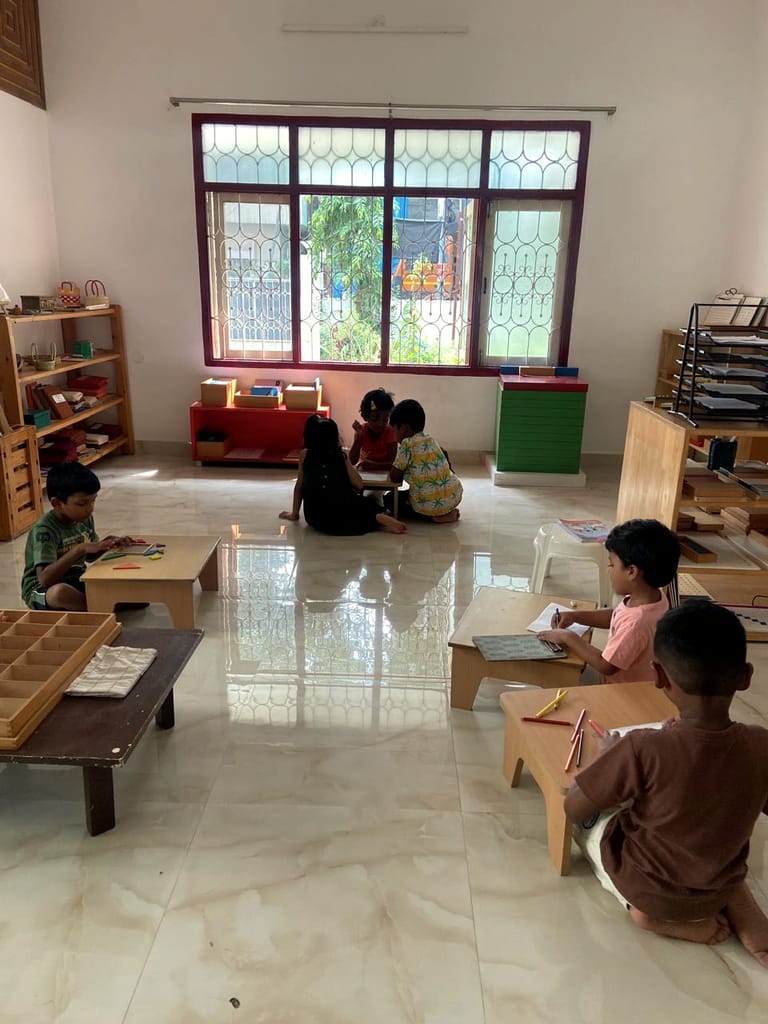 Children working at tables and on the floor, seen from the classroom doorway.