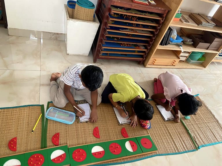 Three children work with red fraction circles and notebooks on floor mats.