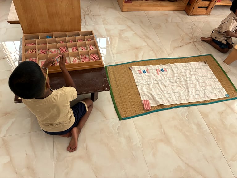 Seen from above, a child chooses pink wooden letters beside a lined cloth on a mat.