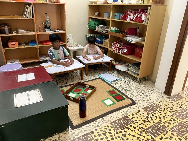 Two children write at a low table among shelves of classroom materials.