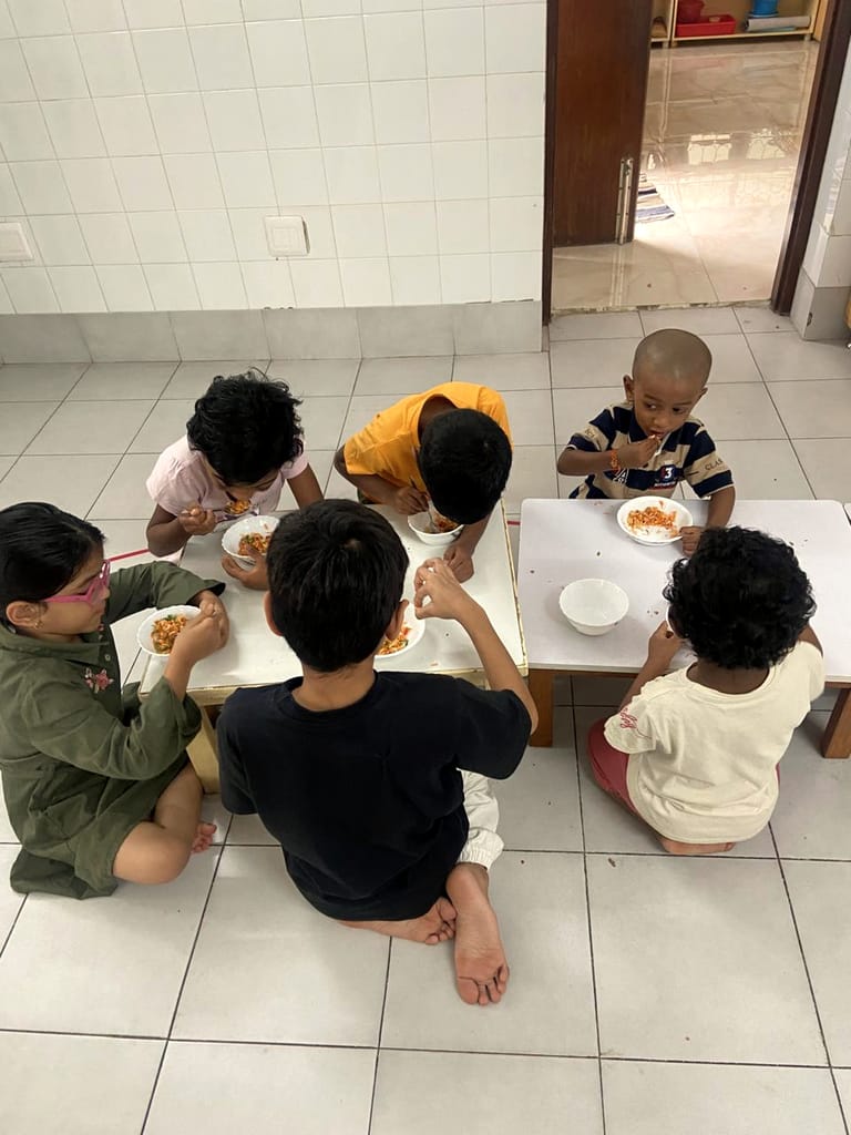 Children sit around two low tables with plates of food.
