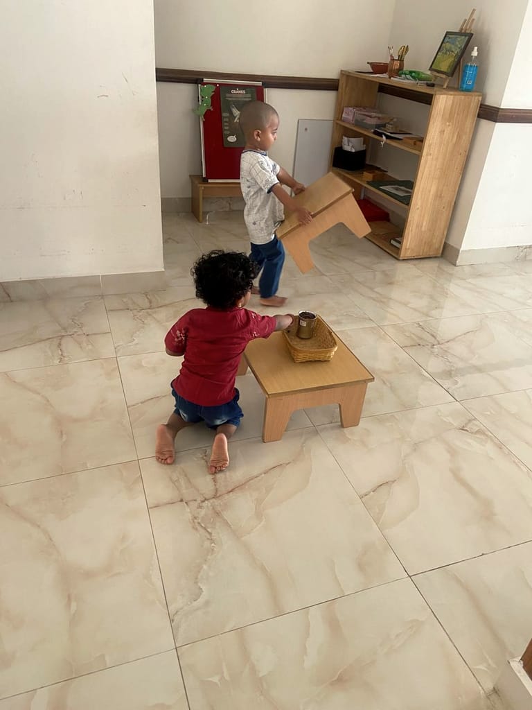 Two young children stand near a small wooden table in the classroom.
